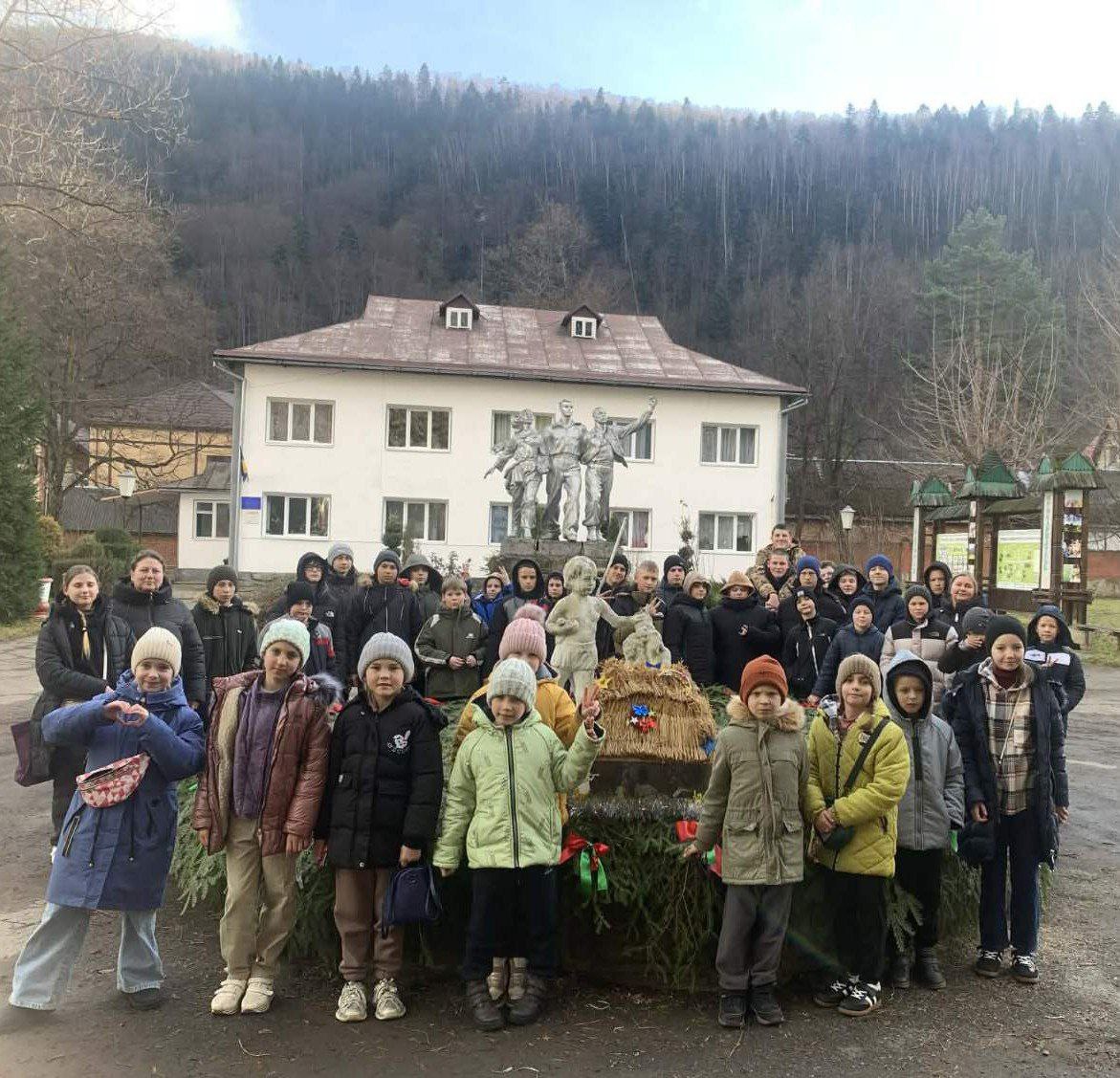 Групова фотографія дорослих і дітей (близько 40 осіб) на відкритому повітрі. Вони стоять перед білою двоповерховою будівлею, на фасаді якої розташована скульптурна композиція. Перед групою знаходиться інсталяція з ялинових гілок та фігурками. На задньому плані видно лісистий схил гори.
