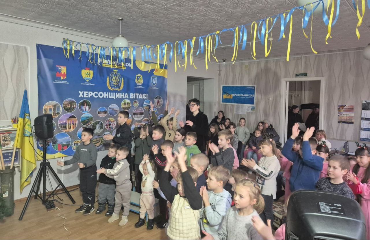 Група дітей та підлітків бере участь у заході в приміщенні, прикрашеному синьо-жовтими стрічками та великим банером із написом: ХЕРСОНЩИНА ВІТАЄ! Близько тридцяти дітей підняли руки вгору, очевидно, реагуючи на ведучого або граючи в гру. На стінах видно плакати та стенд ХЕРСОНСЬКИЙ ХАБ.