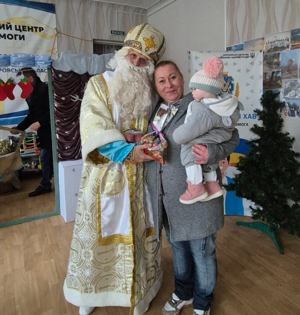 Жінка, що тримає на руках маленьку дитину в рожевій шапці, фотографується із чоловіком у костюмі Святого Миколая (золотий із білим). Святий Миколай вручає їм подарунковий пакунок. На задньому плані видно частину банера Херсонської ОВА та новорічну ялинку.