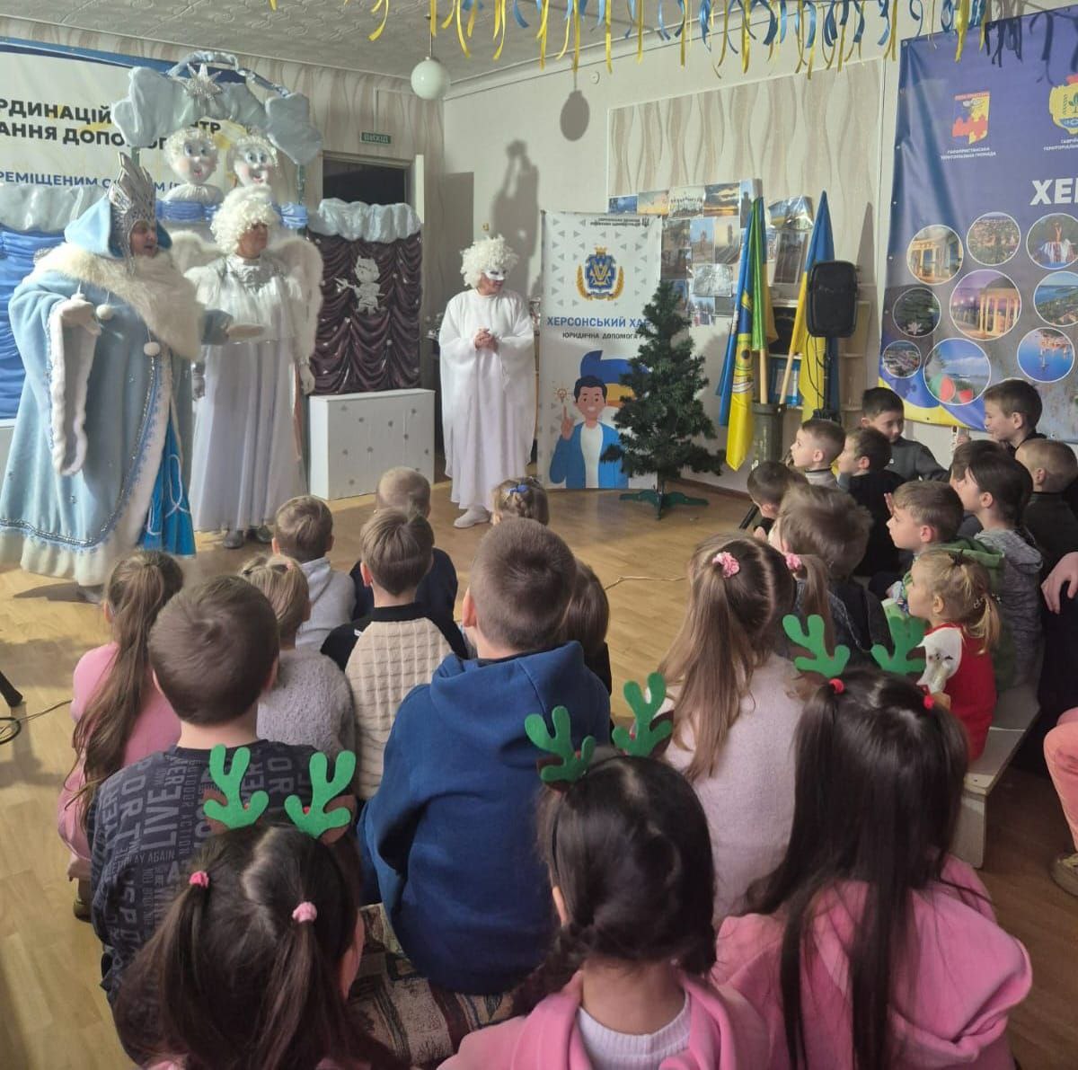 Сцена святкового заходу для дітей у приміщенні Координаційного центру ВПО. Четверо дорослих у костюмах новорічних персонажів (Святого Миколая, Снігової Королеви та Ангелів) стоять на імпровізованій сцені та виступають перед групою дітей, які сидять на підлозі та вдягнені у ріжки оленів. На задньому плані розташований банер з інформацією про Херсонський координаційний центр.