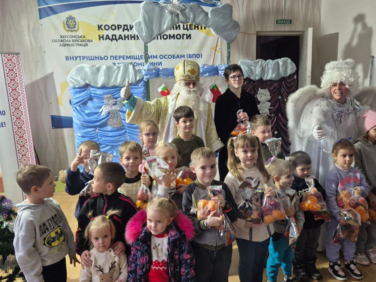Групова фотографія дітей та кількох дорослих, переодягнених у новорічних персонажів (зокрема, Святого Миколая та Снігову Королеву/Ангела), зроблена у приміщенні. Діти тримають у руках святкові пакунки з мандаринами та солодощами. На задньому плані розташований банер з логотипом Херсонської ОВА та написом: КООРДИНАЦІЙНИЙ ЦЕНТР НАДАННЯ ДОПОМОГИ ВНУТРІШНЬО ПЕРЕМІЩЕНИМ ОСОБАМ (ВПО).