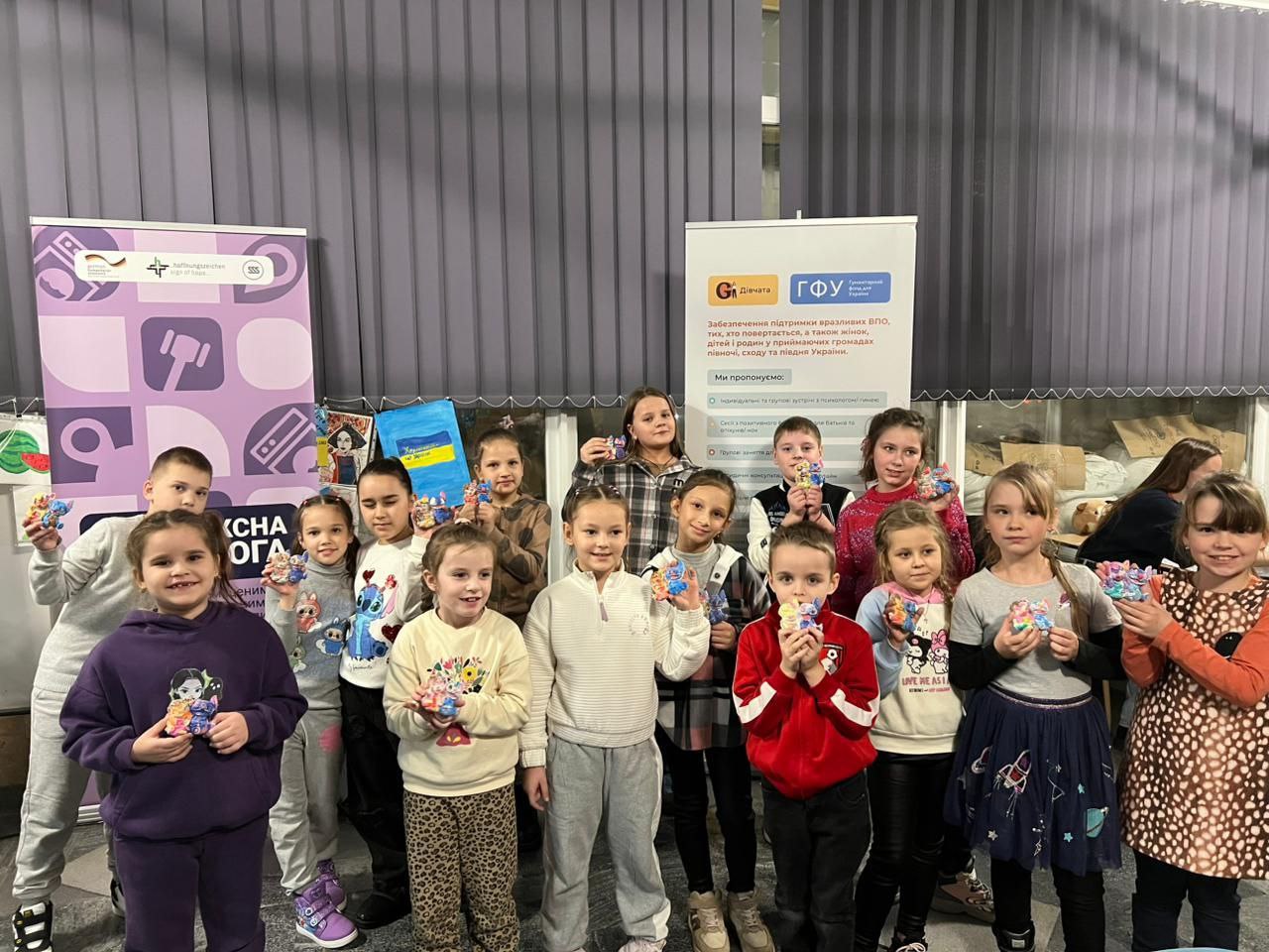 Група з приблизно п'ятнадцяти дітей стоїть в приміщенні на тлі банерів і плакатів, посміхається та демонструє невеликі подарунки, які вони тримають в руках.