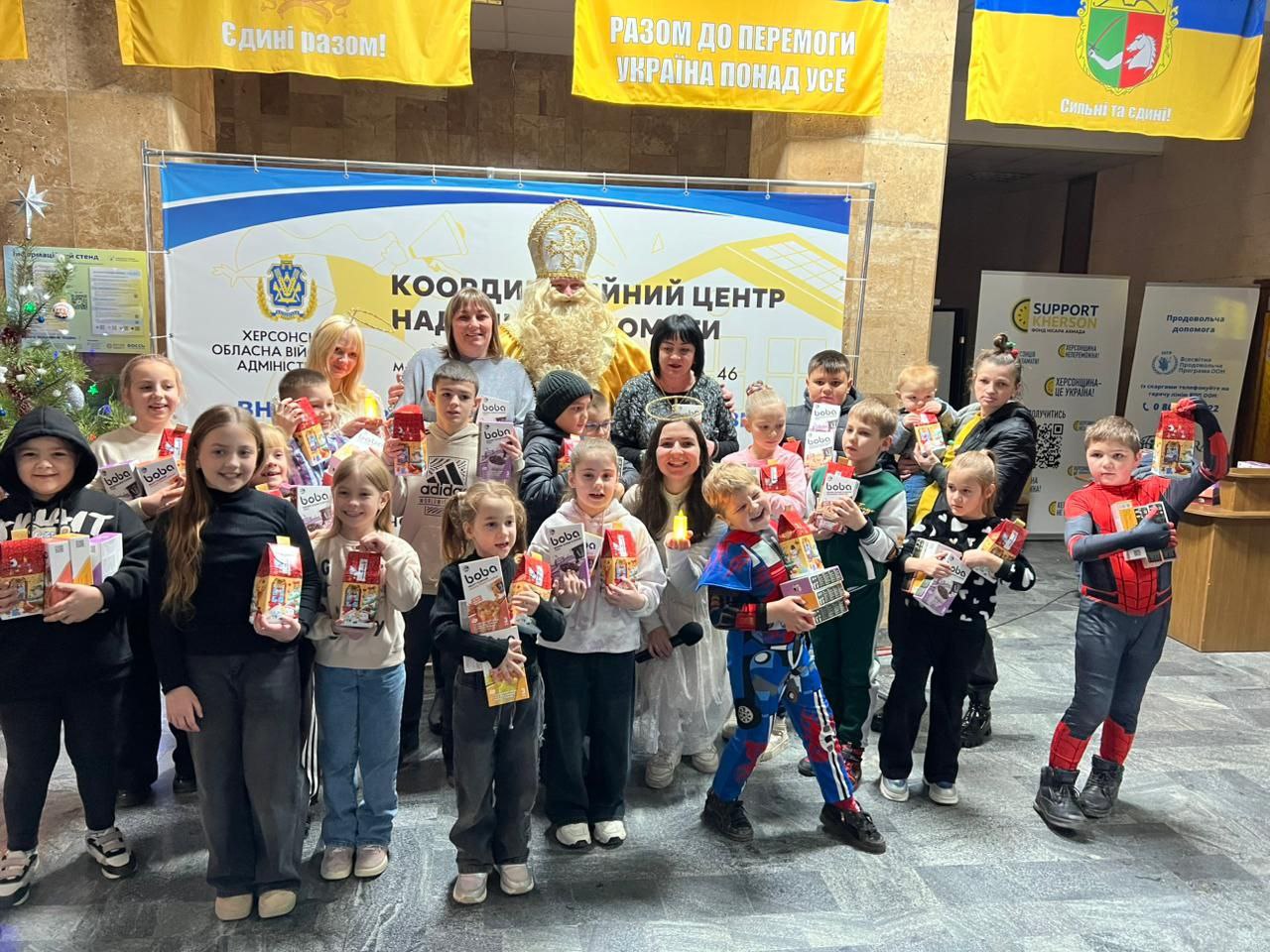 Група дітей, які тримають подарунки, стоїть із двома жінками та чоловіком, одягненим як Святий Миколай, на тлі банера КООРДИНАЦІЙНИЙ ЦЕНТР та українських прапорів.
