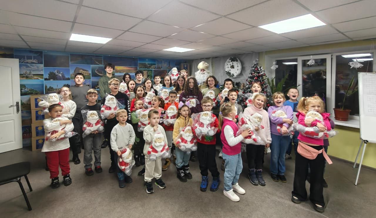 Група дітей та підлітків, які радісно тримають м'які іграшки у вигляді Святих Миколайчиків, стоять у приміщенні з дорослим чоловіком у костюмі Святого Миколая та прикрашеною ялинкою на задньому плані.