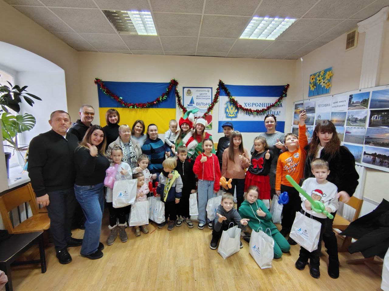 Група людей, включаючи дорослих та багато дітей, стоять разом у приміщенні з двома великими прапорами України та Херсонщини на стіні. У центрі доросла жінка, одягнена як Святий Миколай або Снігуронька, посміхається. Діти тримають білі подарункові пакети та повітряні кульки. Це виглядає як святковий захід.