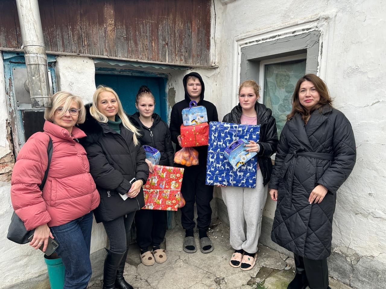 Група з шести людей (три дорослі жінки та троє підлітків) стоїть на вулиці біля старої побіленої будівлі, тримаючи різноманітні святково упаковані подарунки та фрукти.