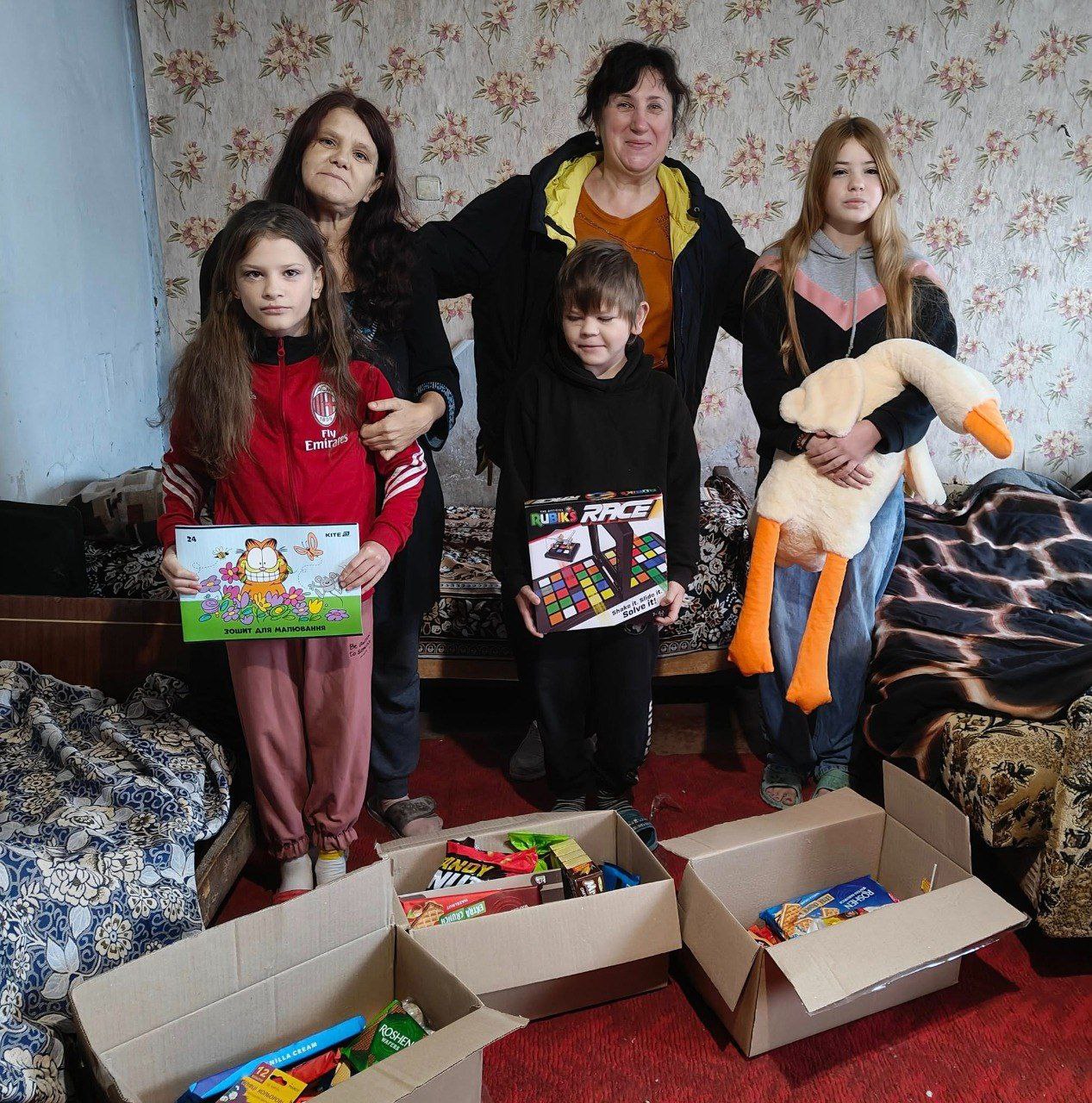 Група з п'яти людей (дві дорослі жінки та троє дітей) стоїть у кімнаті, тримаючи іграшки та коробки з подарунками, а також три відкриті коробки з цукерками та солодощами стоять на підлозі перед ними.