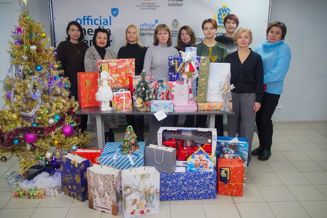 Група з дев'яти жінок стоїть біля столу, заваленого красиво упакованими новорічними та різдвяними подарунками, поруч стоїть прикрашена ялинка.