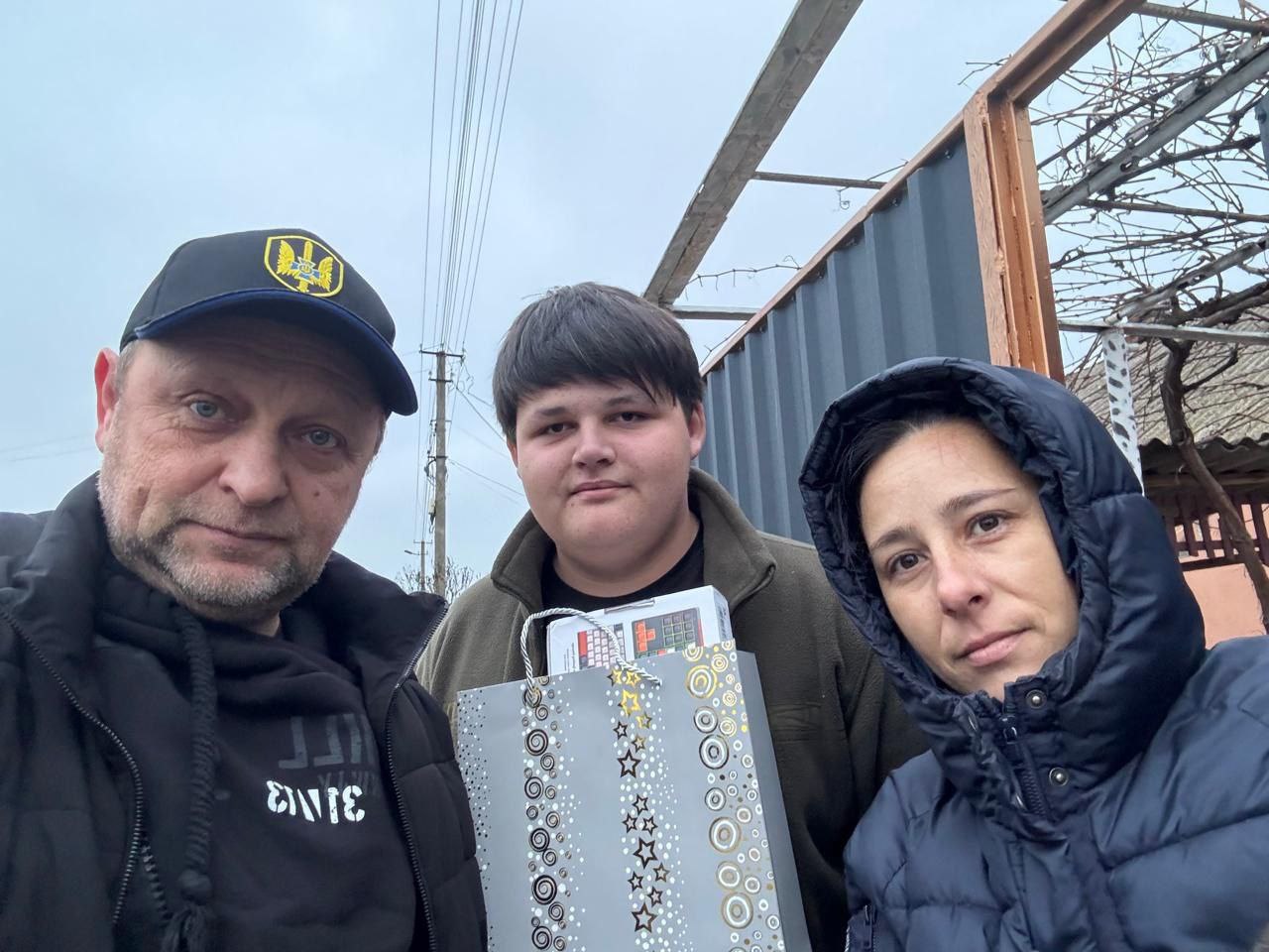 Селфі трьох людей – чоловіка у кепці з емблемою, молодого хлопця, який тримає подарунковий пакет, та жінки у капюшоні – на тлі подвір'я та похмурого неба.