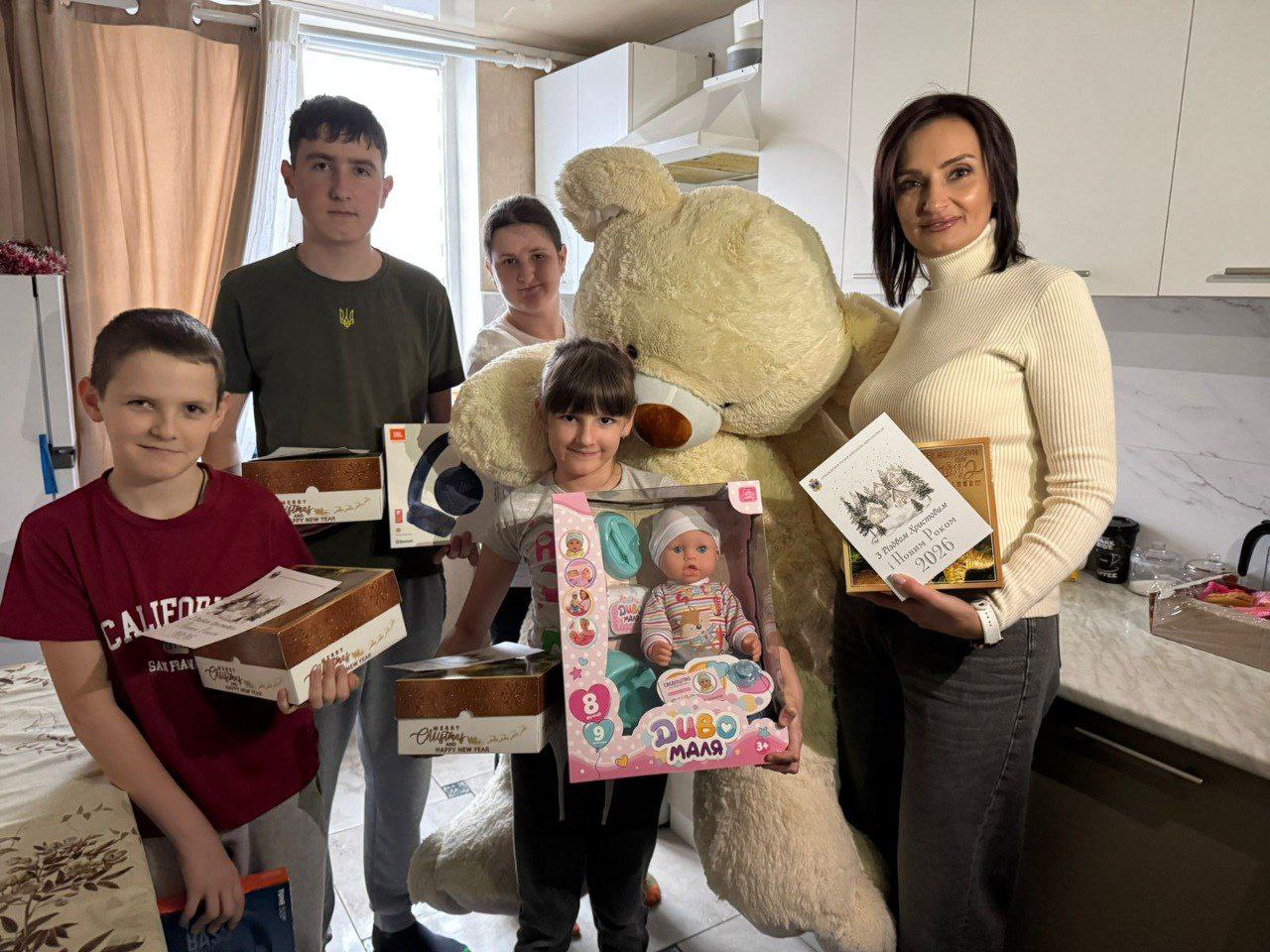 П'ятеро людей, включаючи жінку, яка тримає коробку, і чотирьох дітей різного віку, які тримають подарунки (коробки, ляльку), позують у приміщенні поруч із великим плюшевим ведмедем.