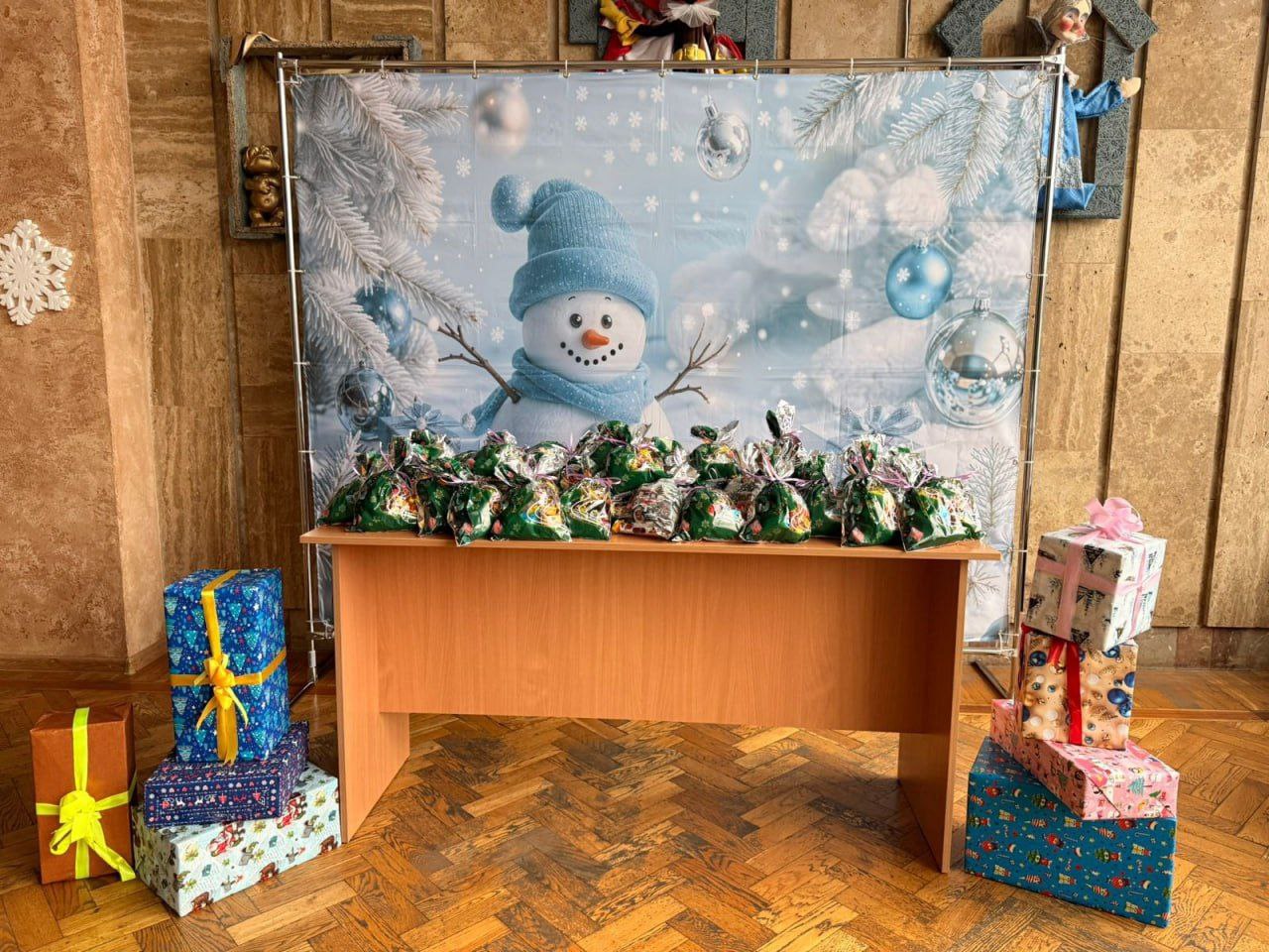 На столі, прикрашеному новорічним задником зі сніговиком, розкладені численні невеликі зелені мішечки з подарунками, а також навколо столу стоять великі святково упаковані коробки.