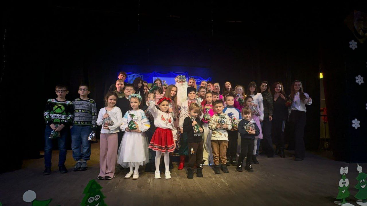 Групова фотографія дітей та дорослих на сцені після вистави. У центрі стоїть чоловік у костюмі Святого Миколая. Більшість дітей тримають у руках подарунки, деякі дівчата одягнені у вишиванки та народні костюми. На задньому плані видно український прапор та герб, а по боках сцени — декорації у вигляді ялинок та сніжинок.