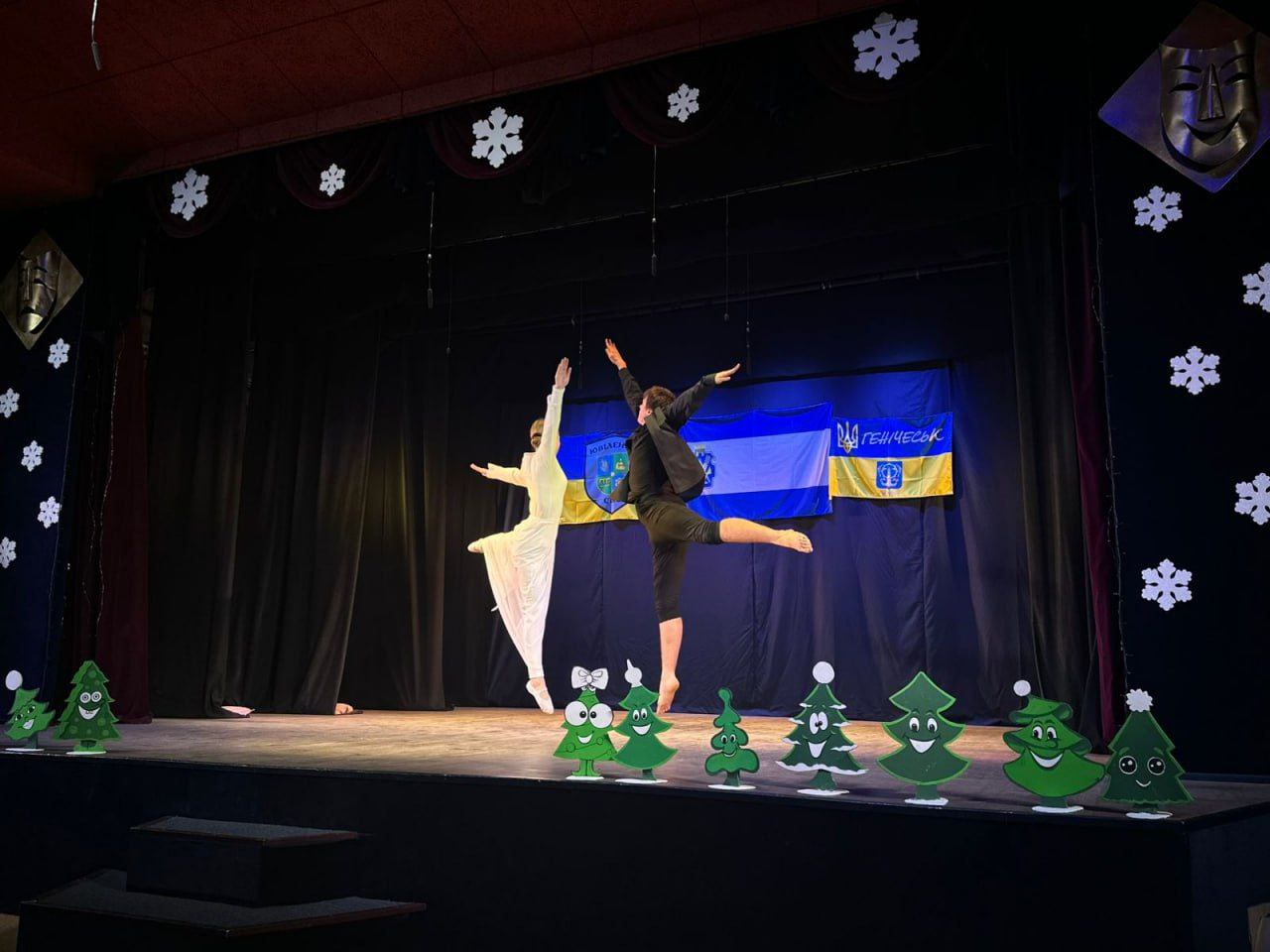 На сцені, прикрашеній різдвяними сніжинками та вирізаними ялинками, двоє танцюристів виконують балетний елемент на тлі синьо-жовтого прапора з емблемою Генічеська.