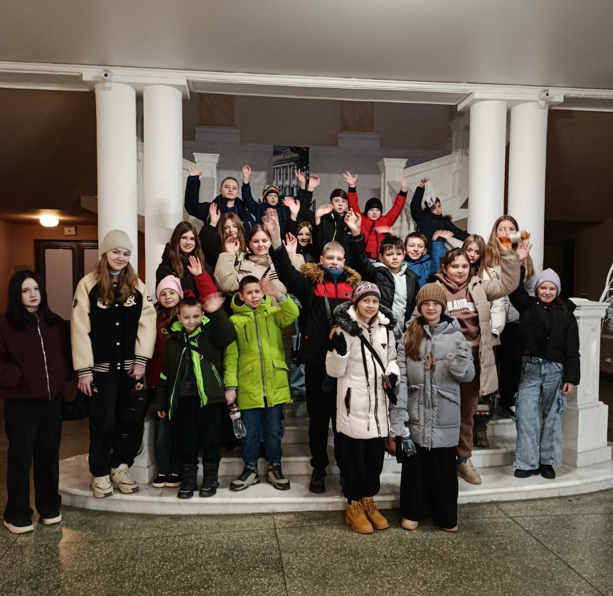 Групове фото, зроблене у приміщенні. Приблизно двадцять дітей та підлітків різного віку стоять на широких мармурових сходах з білими колонами, багато хто з них махає руками.