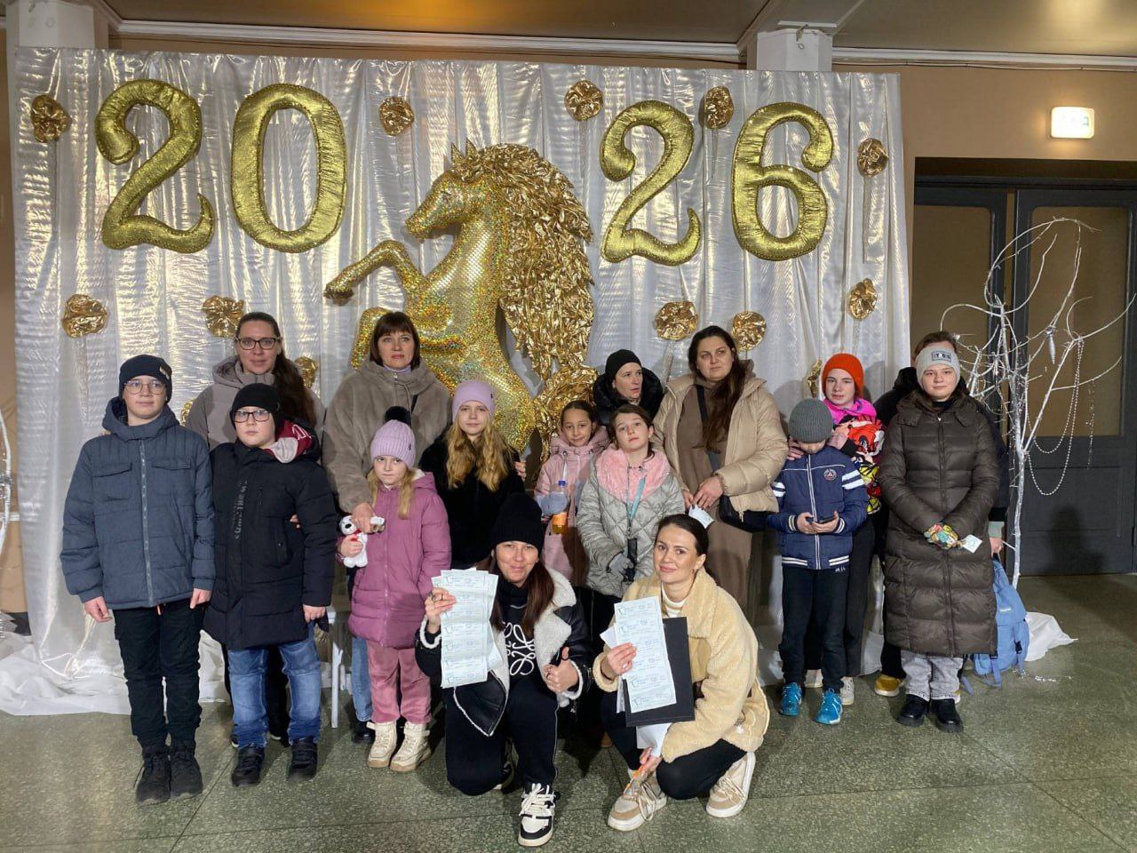 Груповий портрет дорослих і дітей, які стоять перед святковим фотофоном із великими золотими повітряними кульками у формі чисел 2026 та великим зображенням коня. Дві жінки на передньому плані сидять навпочіпки і тримають аркуші паперу.