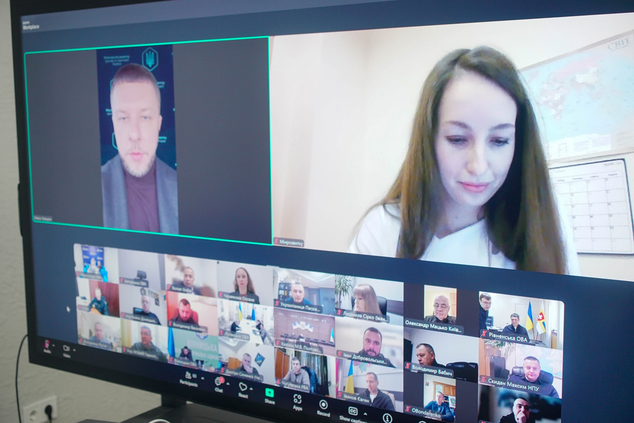 Знімок екрана комп'ютера, що відображає відеоконференцію в Zoom. У верхньому ряду показано двох основних спікерів: чоловіка з бородою в бордовому светрі (зліва) та жінку з довгим темним волоссям (справа). Нижче — галерея з маленькими віконцями, де видно багато інших учасників, включно з особами, підписаними як: Херсонська ОВА та іншими.