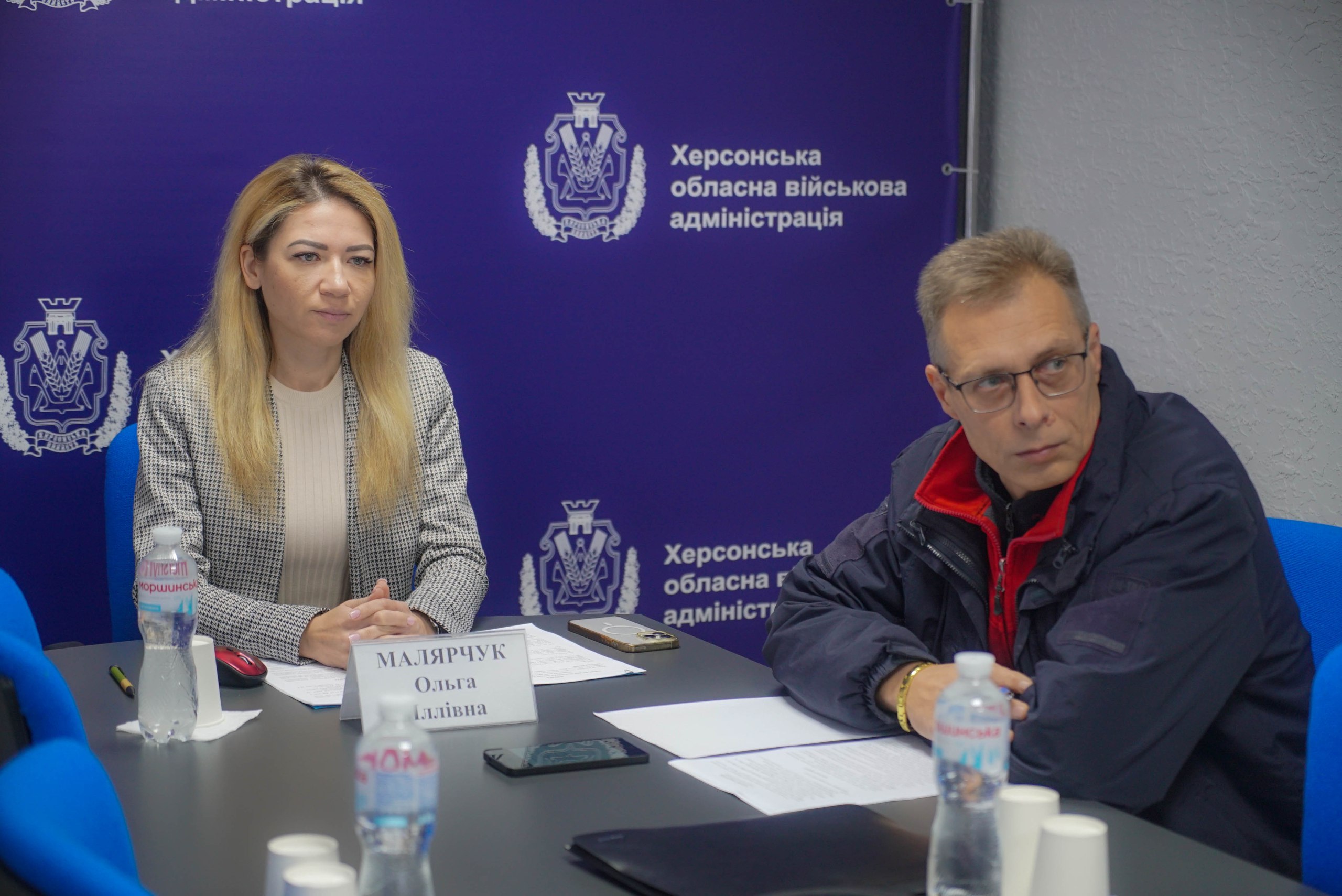 Жінка та чоловік сидять за столом під час пресконференції. На столі перед жінкою стоїть табличка з написом. На задньому плані — синій банер із зображенням емблеми та написом: Херсонська обласна військова адміністрація.