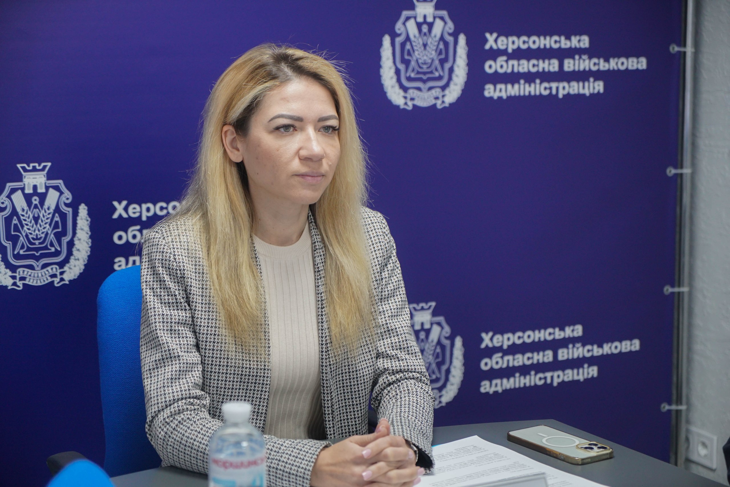 Жінка зі світлим волоссям сидить за столом у піджаку в клітинку перед синім фоном з емблемою та написом: Херсонська обласна військова адміністрація.