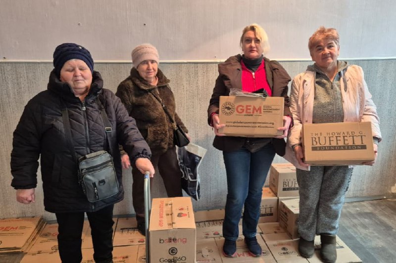 Чотири жінки стоять у приміщенні серед коробок з гуманітарною допомогою. Дві жінки праворуч тримають картонні коробки від GEM Global Empowerment Mission та The Howard G. Buffett Foundation. На підлозі видно багато коробок, зокрема з логотипами Good360.