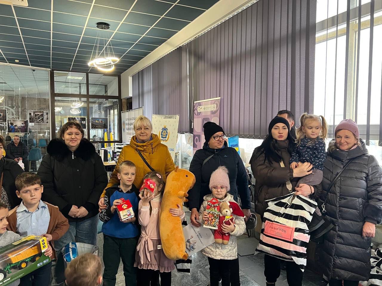 Група дорослих та дітей стоять у холі біля великого вікна. Одна жінка тримає маленьку дівчинку та смугастий пакет, а інші дорослі стоять на передньому плані разом з дітьми, які тримають іграшки та подарунки, такі як плюшеві іграшки, машинки та книжки. На задньому плані видно рекламні банери.