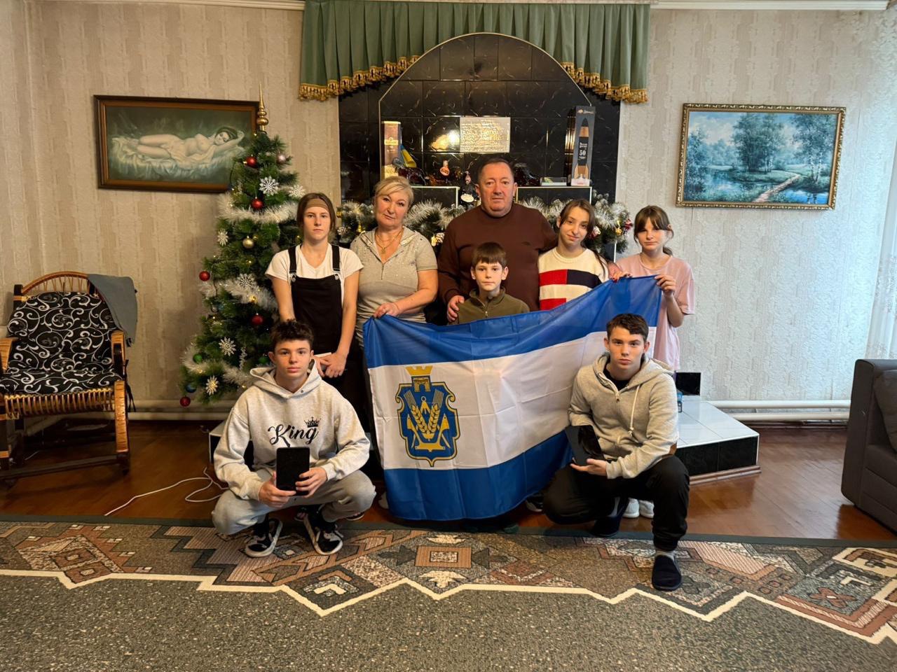 Груповий портрет: чоловік та жінка середнього віку стоять у центрі, оточені шістьма дітьми та підлітками (троє хлопчиків, троє дівчаток). Вони тримають розгорнутий біло-блакитний прапор із гербом міста Херсона. На задньому плані видно новорічну ялинку та камін.