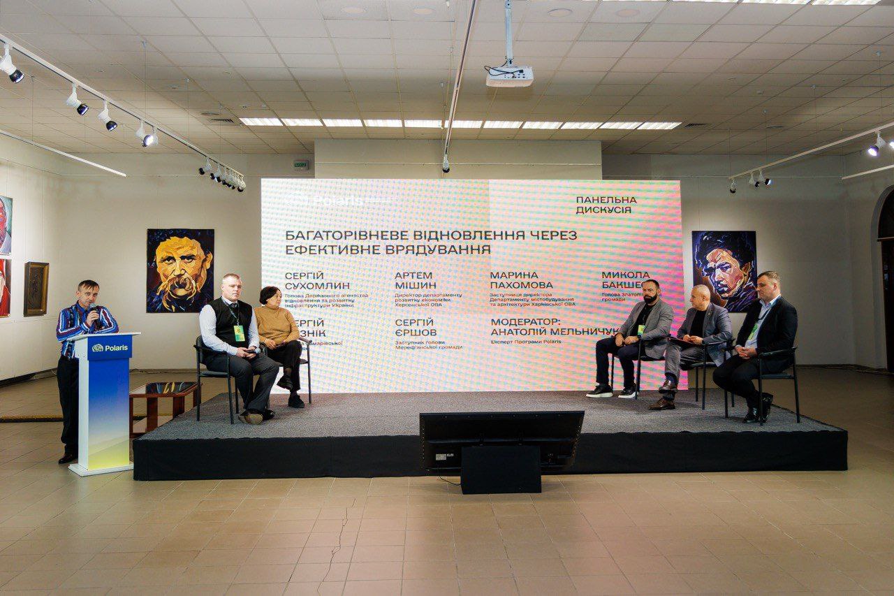 Панельна дискусія на сцені, де п'ять людей сидять у кріслах, а один чоловік стоїть за трибуною ліворуч. На великому екрані позаду них великими літерами написано: БАГАТОРІВНЕВЕ ВІДНОВЛЕННЯ ЧЕРЕЗ ЕФЕКТИВНЕ ВРЯДУВАННЯ та ПАНЕЛЬНА ДИСКУСІЯ.