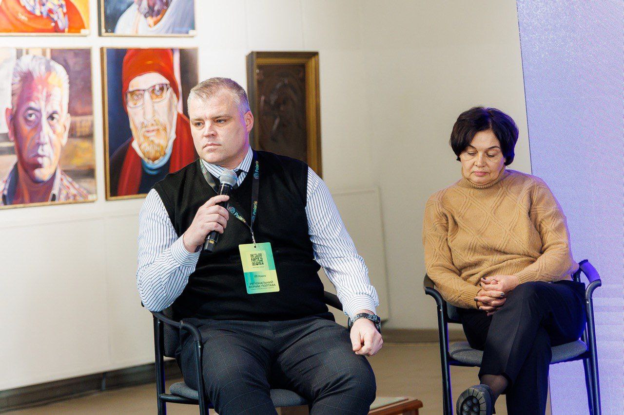 Чоловік у смугастій сорочці та чорному жилеті сидить на стільці, тримаючи мікрофон, і дивиться перед собою. Поруч сидить жінка у світло-коричневому светрі. На задньому плані — картини з портретами.