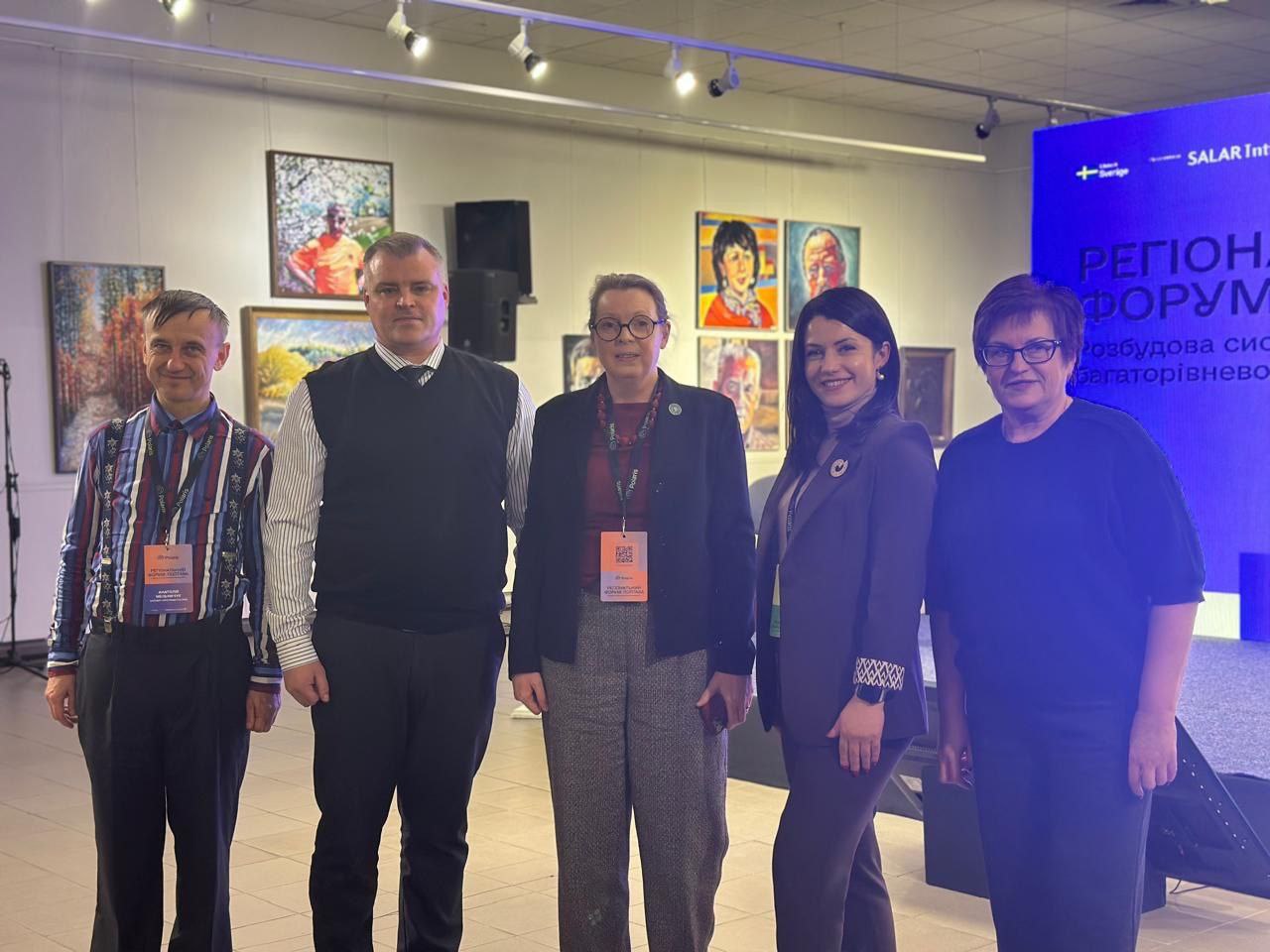 П'ятеро людей, троє жінок і двоє чоловіків, стоять поруч і дивляться в камеру на виставці. На задньому плані видно картини та банер.