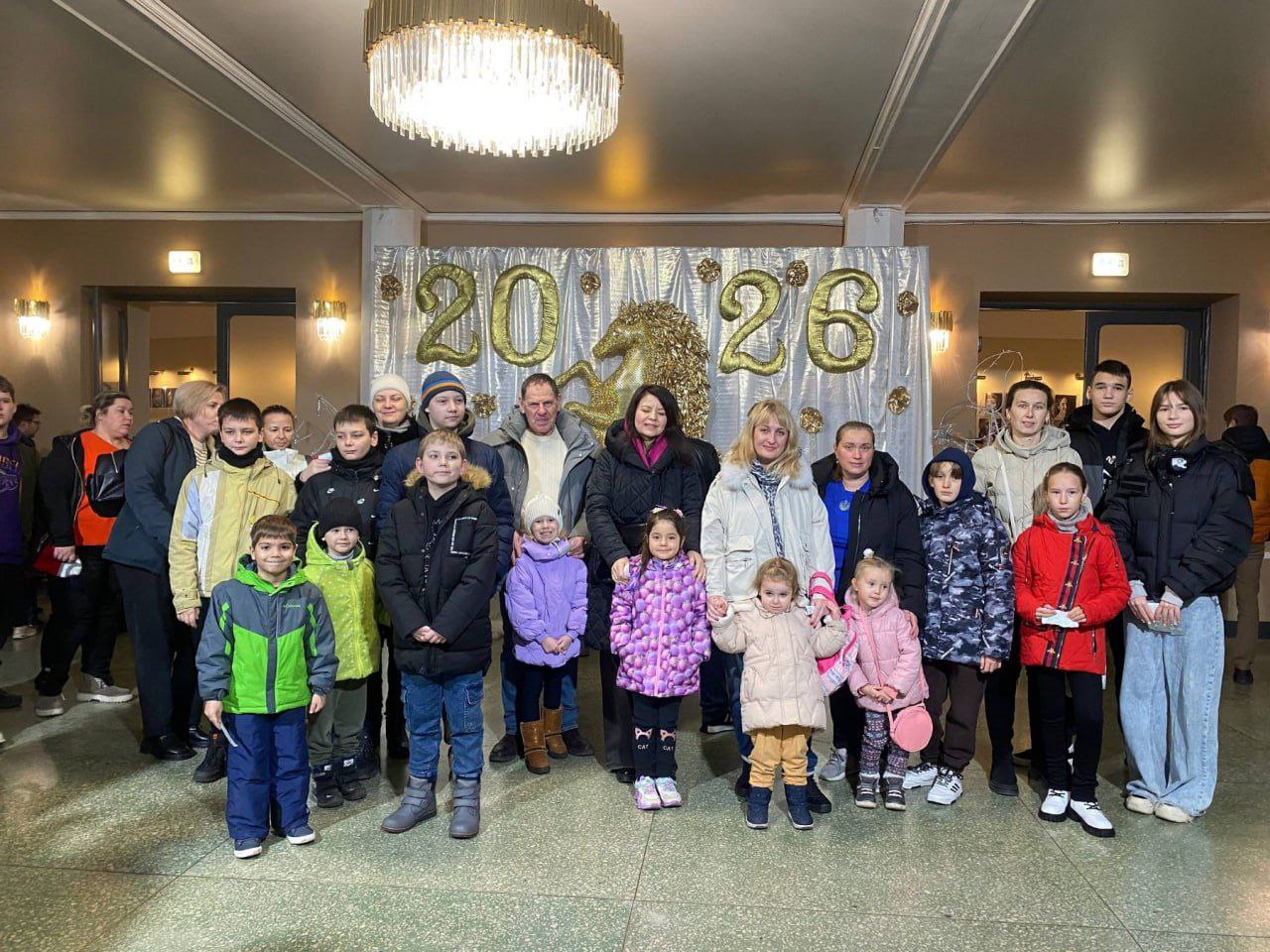 Групове фото близько двадцяти осіб, включно з дорослими та дітьми, які стоять у великому холі. На задньому плані видно великі золоті повітряні кульки у формі чисел 2026 та великим зображенням коня на блискучому тлі, а також масивну люстру.