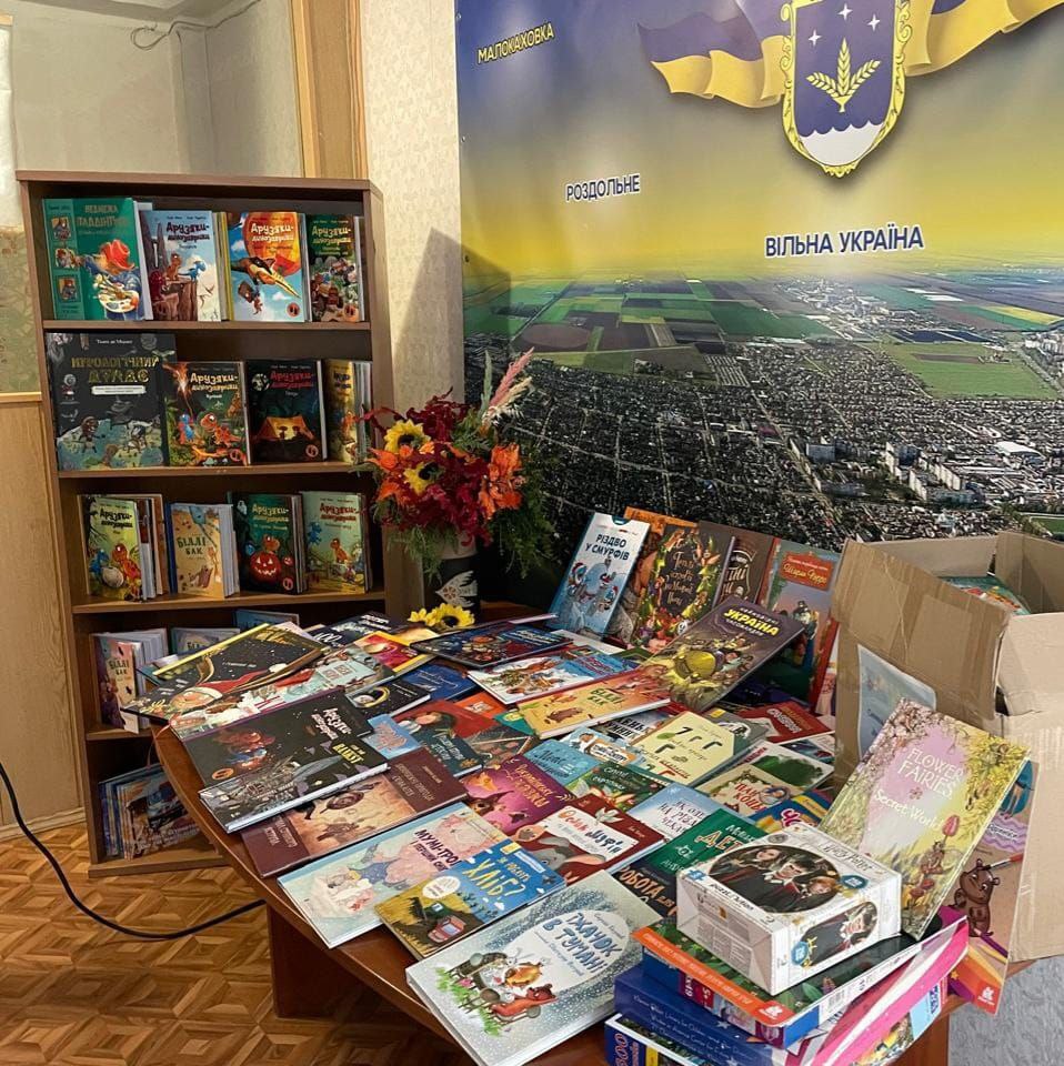 Стіл, заповнений розгорнутими та складеними дитячими книгами, ігровими коробками та букетом квітів, на тлі книжкової шафи та великого банера Каховської міської територіальної громади.