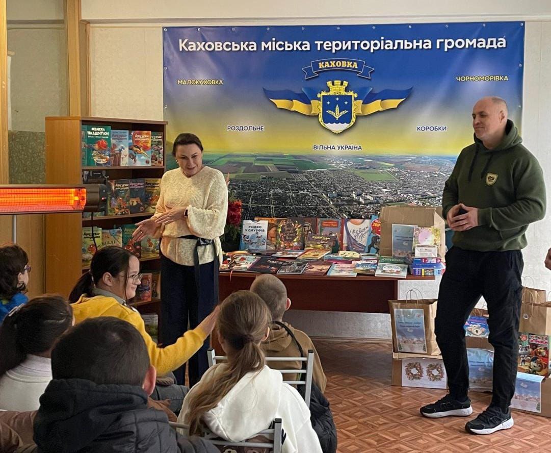 Жінка виступає перед групою дітей, що сидять, поруч стоїть чоловік. На задньому плані банер Каховської міської територіальної громади та стіл з книгами.