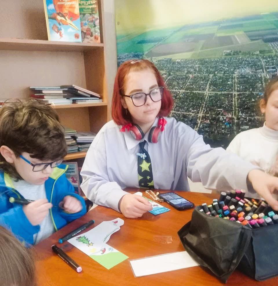 Дівчина-підліток у чорних окулярах і з червоним волоссям сидить за столом поруч із молодшим хлопчиком. На столі лежать маркери та мобільний телефон, діти зайняті малюванням чи творчістю.