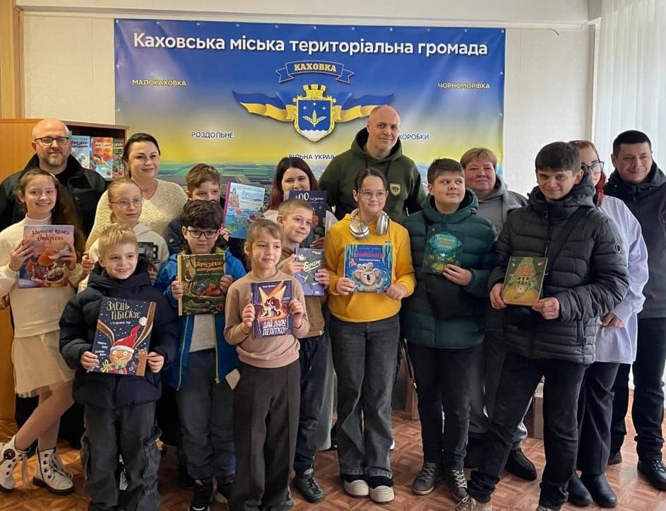 Групове фото дорослих і дітей, які тримають у руках книги, на тлі банера Каховської міської територіальної громади.