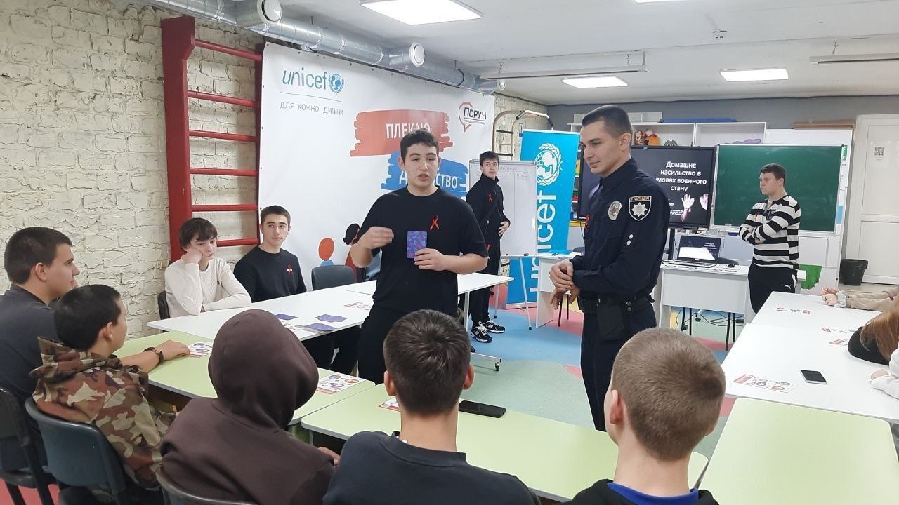 Група підлітків сидить за столами, уважно слухаючи, як хлопець у чорній футболці жестикулює, стоячи поруч із поліцейським у формі. На задньому плані банер UNICEF та червона пожежна драбина.