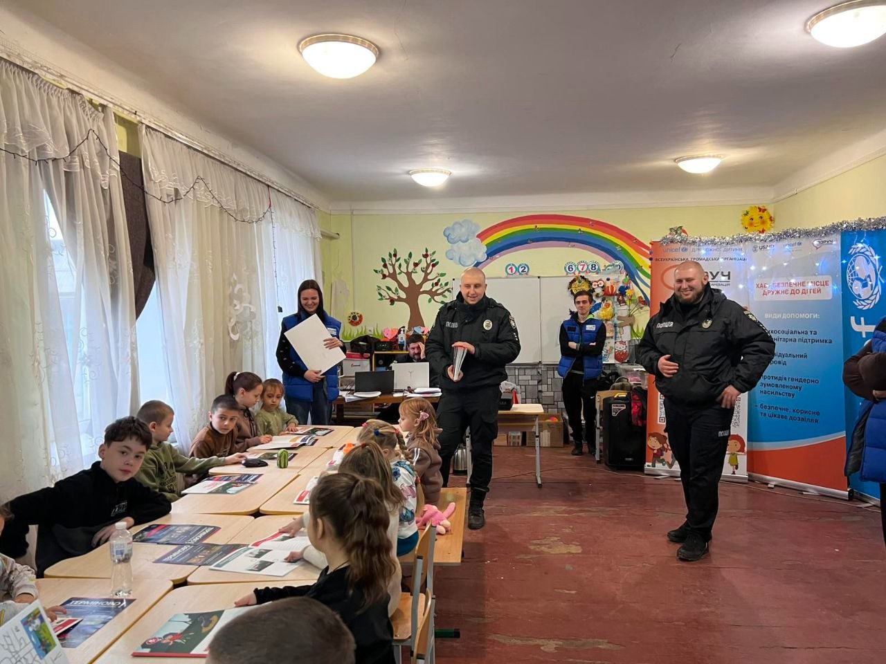 Два поліцейські (один тримає книгу), двоє волонтерів (молодий чоловік, молода жінка) та група маленьких дітей, що сидять за столами, у кімнаті з декором у вигляді веселки та банером Поруч. Каховська громада.
