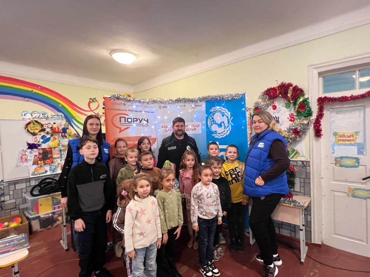 Групове фото, де стоять декілька дорослих (включно з поліцейським) та багато дітей, на тлі святкового декору та банерів UNICEF і Поруч. Каховська громада.