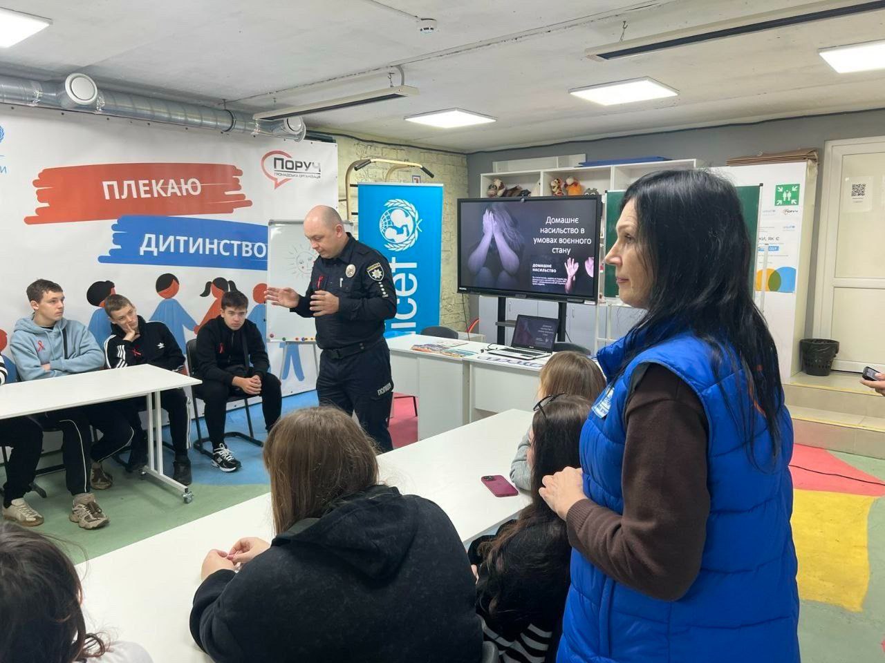 Поліцейський у формі стоїть і активно спілкується з підлітками, що сидять за столами, під час заходу, присвяченого темі: Домашнє насильство в умовах воєнного стану, що відображено на екрані позаду. Молода жінка-волонтер стоїть позаду.