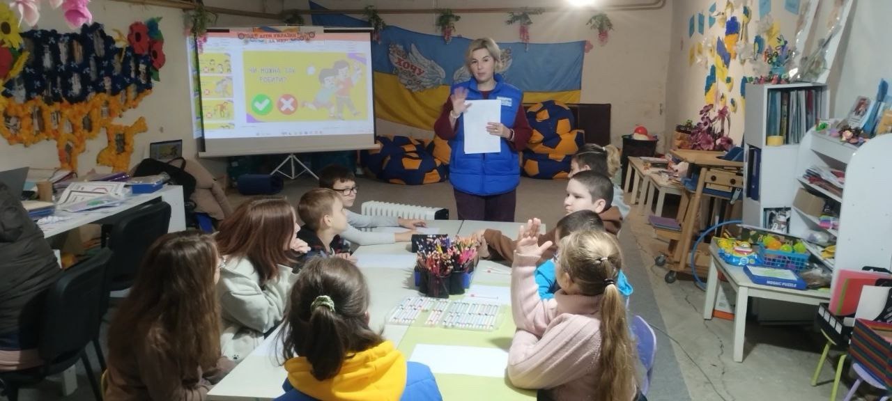 Жінка в синьому жилеті проводить заняття для групи дітей у підвальному приміщенні (бомбосховищі), використовуючи проектор та плакати. Діти сидять за столом і активно піднімають руки.