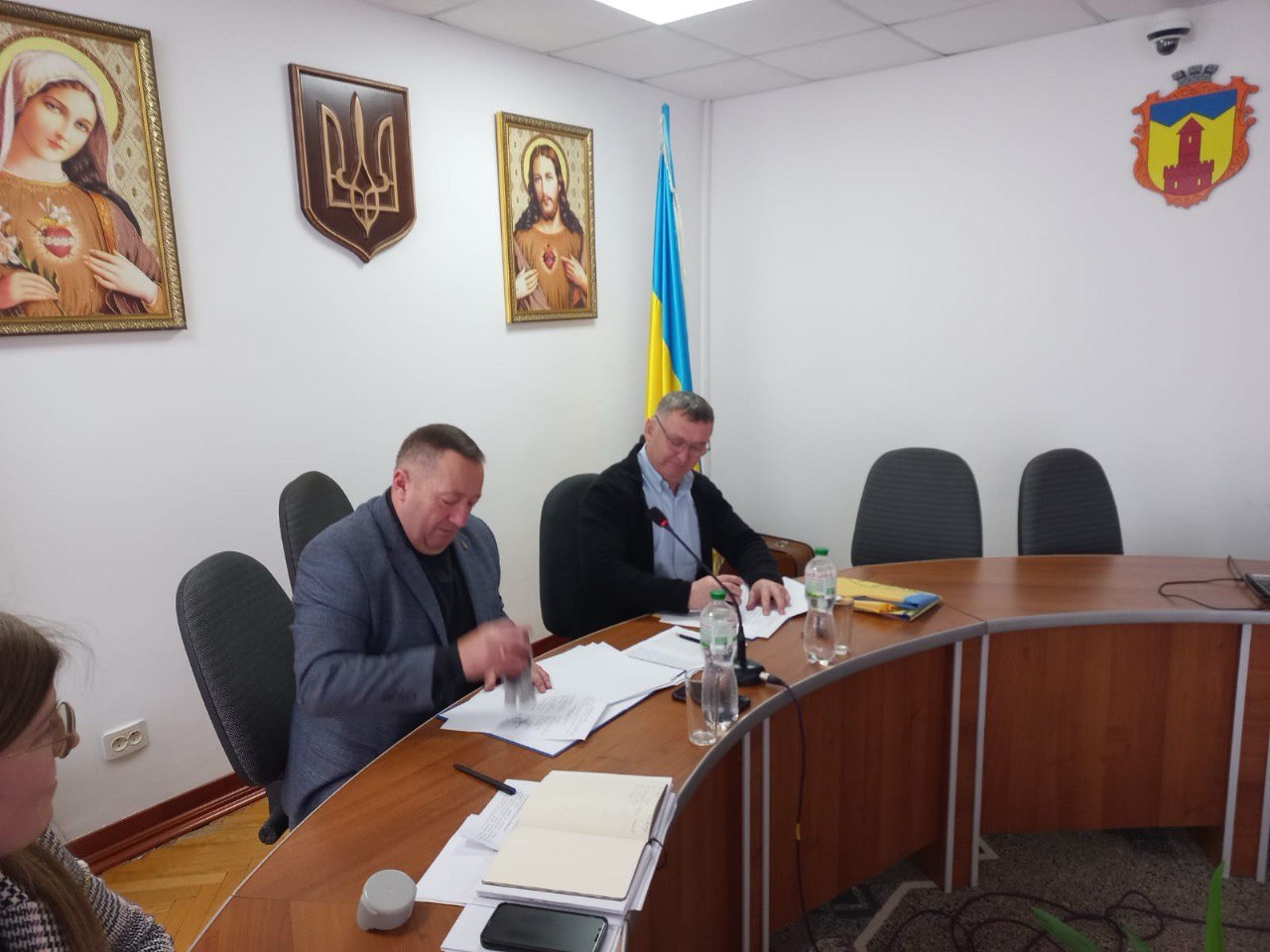 Два чоловіки сидять за великим столом у конференц-залі, працюючи з документами. На стіні за ними висять ікони, герб України та український прапор.