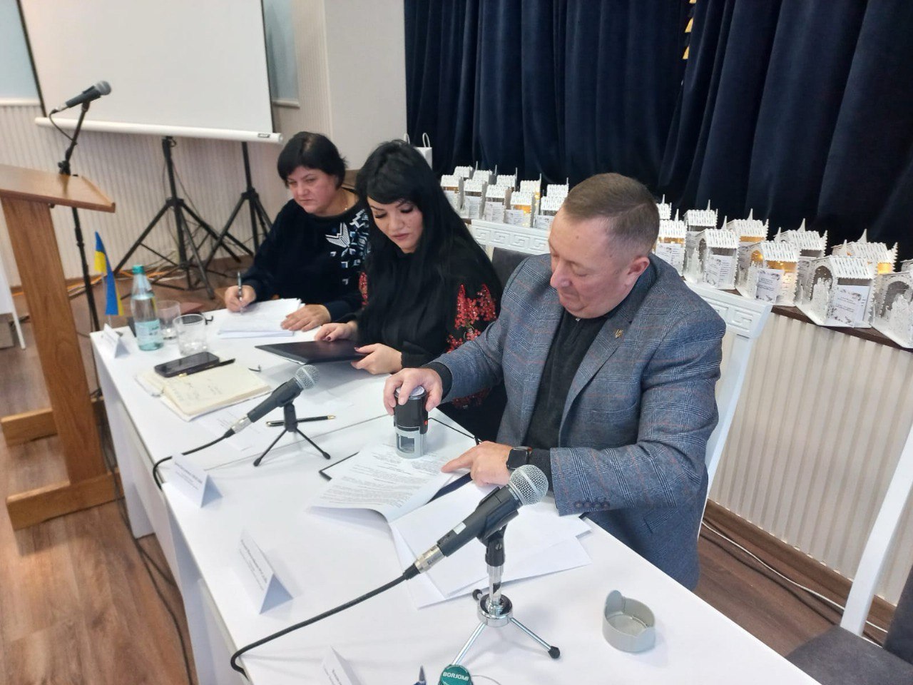 Група з трьох осіб (дві жінки та чоловік у піджаку) сидять за столом під час офіційного заходу, підписуючи та штампуючи документи.