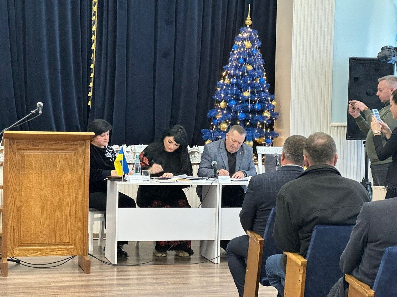 Три особи (дві жінки та чоловік) сидять за столом, підписуючи документи під час офіційного заходу. На задньому плані видно велику новорічну ялинку, прикрашену синіми та золотими вогниками, а також темні штори. На передньому плані видно спини слухачів.