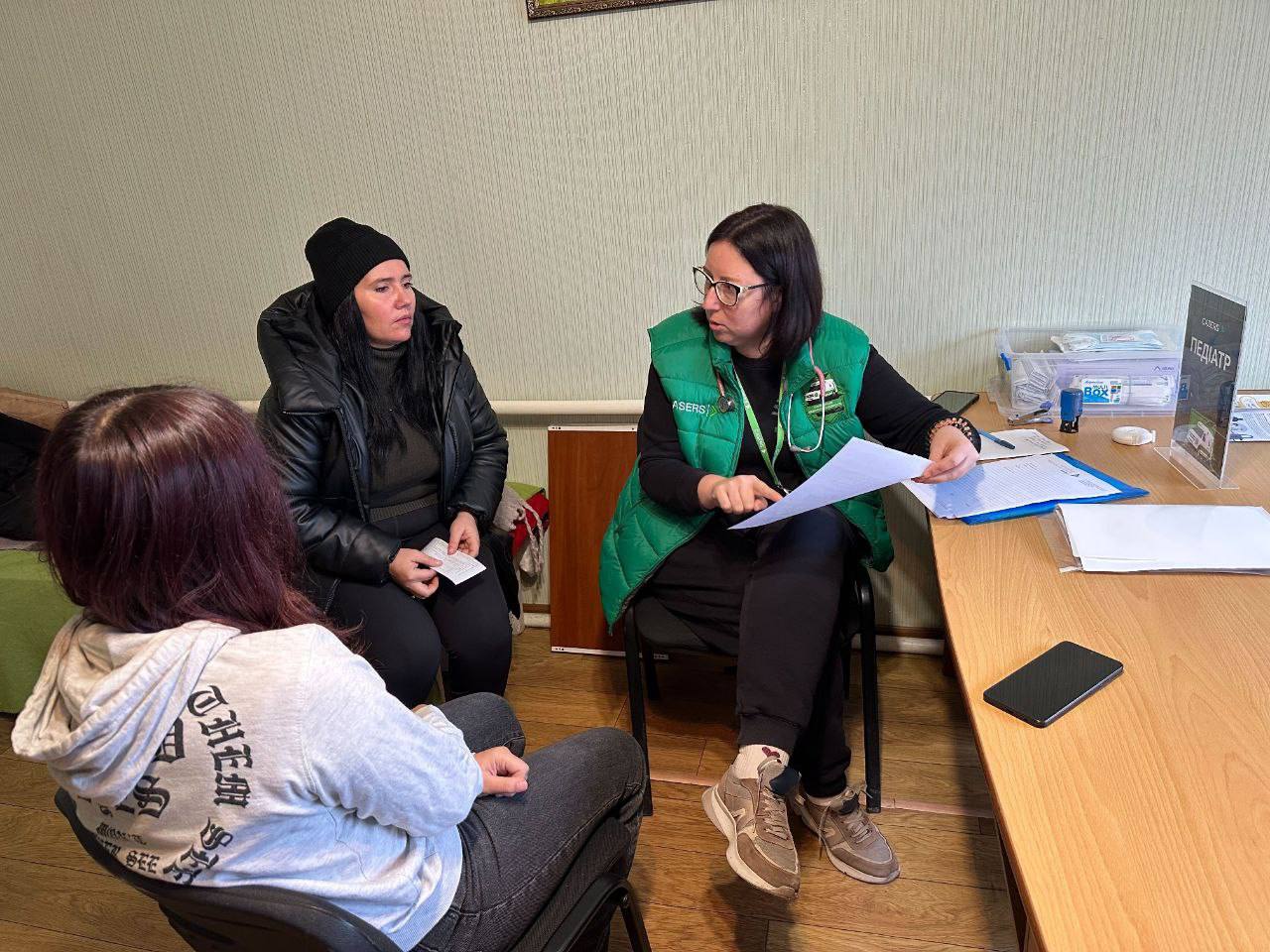 Жінка та дівчина, одягнені в зимовий одяг, сидять навпроти лікарки-педиатрині, яка сидить за столом і тримає в руках документи, в приміщенні схожому на медичний кабінет.