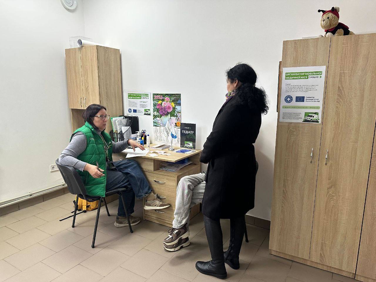 Лікарка-педиатриня в зеленому жилеті сидить за робочим столом, консультуючи жінку в темному пальто, яка стоїть, та людину, що сидить перед столом, у приміщенні схожому на медичний кабінет.