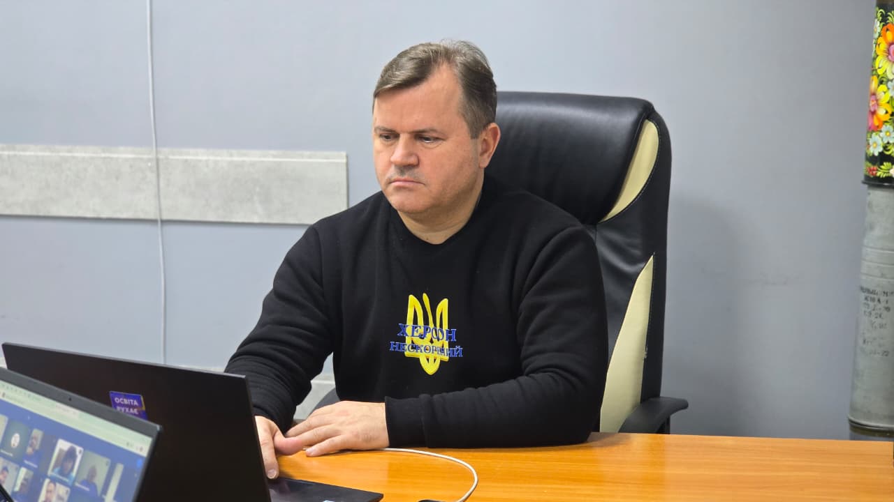Чоловік у чорній кофті з українським гербом сидить за столом, працюючи за ноутбуком і беручи участь у відеоконференції.