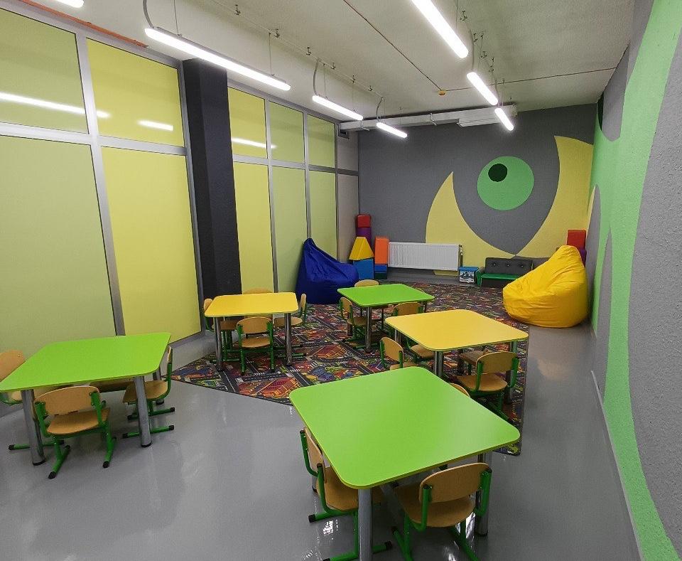 Яскрава дитяча кімната із зеленими та жовтими столиками, маленькими стільцями, килимом із візерунком та великими м'якими кріслами-мішками на тлі жовто-сірих стін.
