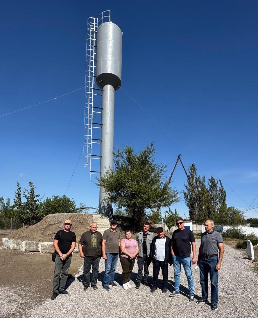 Група з восьми людей (семеро чоловіків та одна жінка) стоїть перед високою сірою водонапірною вежею на тлі блакитного неба та насипу ґрунту.
