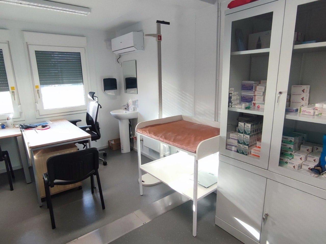 Медичний кабінет із білою шафою, повною ліків, оглядовим столиком для немовлят із ростоміром, умивальником та офісним столом біля вікна.