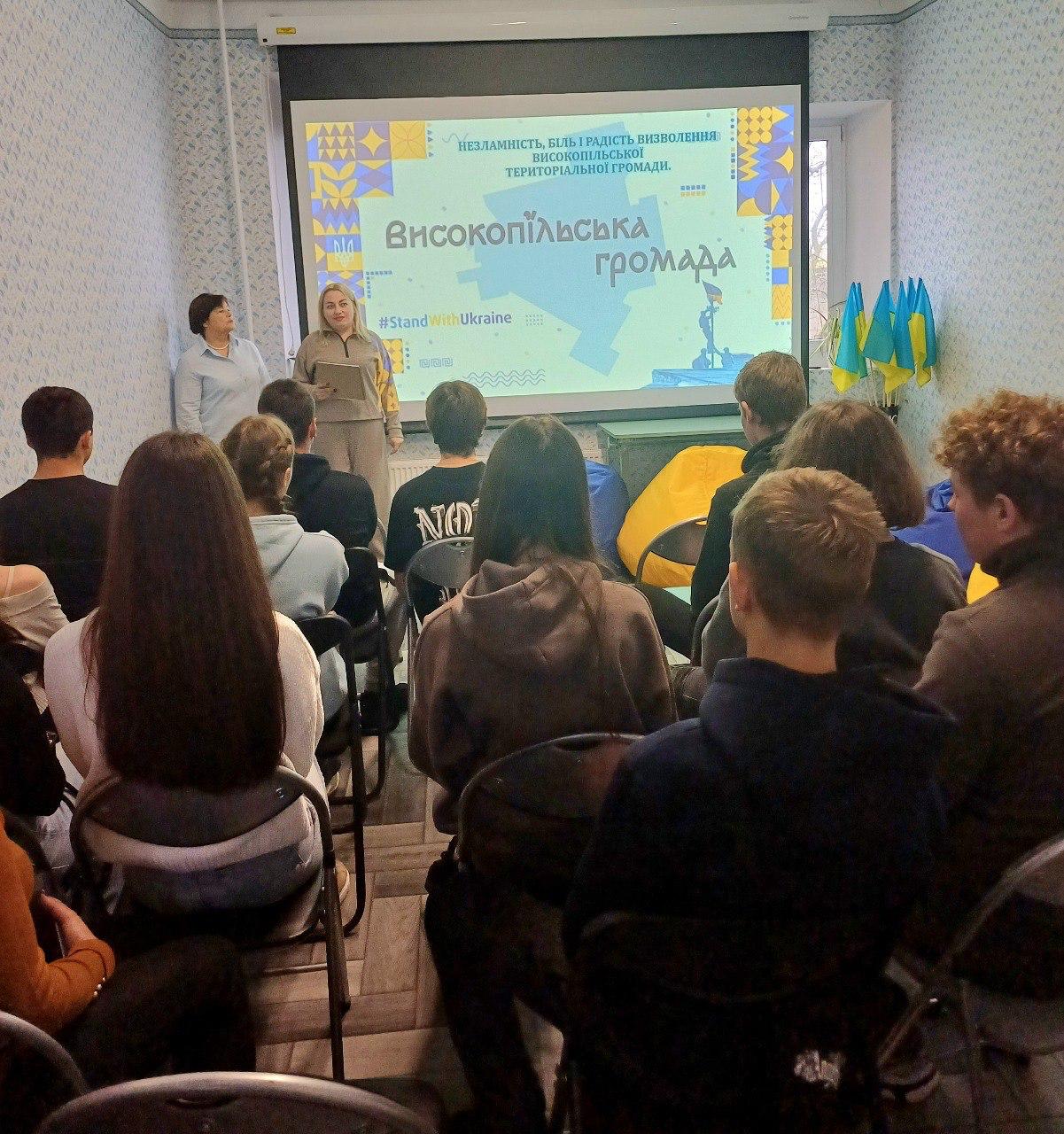 Дві жінки проводять презентацію для групи молоді в аудиторії, на проекційному екрані напис: Високопільська громада та хештег #StandWithUkraine.