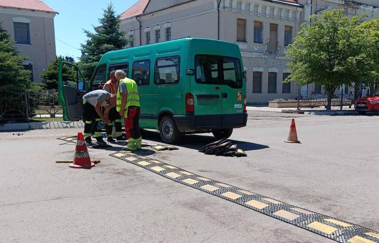 Троє робітників у світловідбивних жилетах встановлюють лежачий поліцейський (штучну нерівність) на дорозі поруч із зеленим мікроавтобусом у міському середовищі.