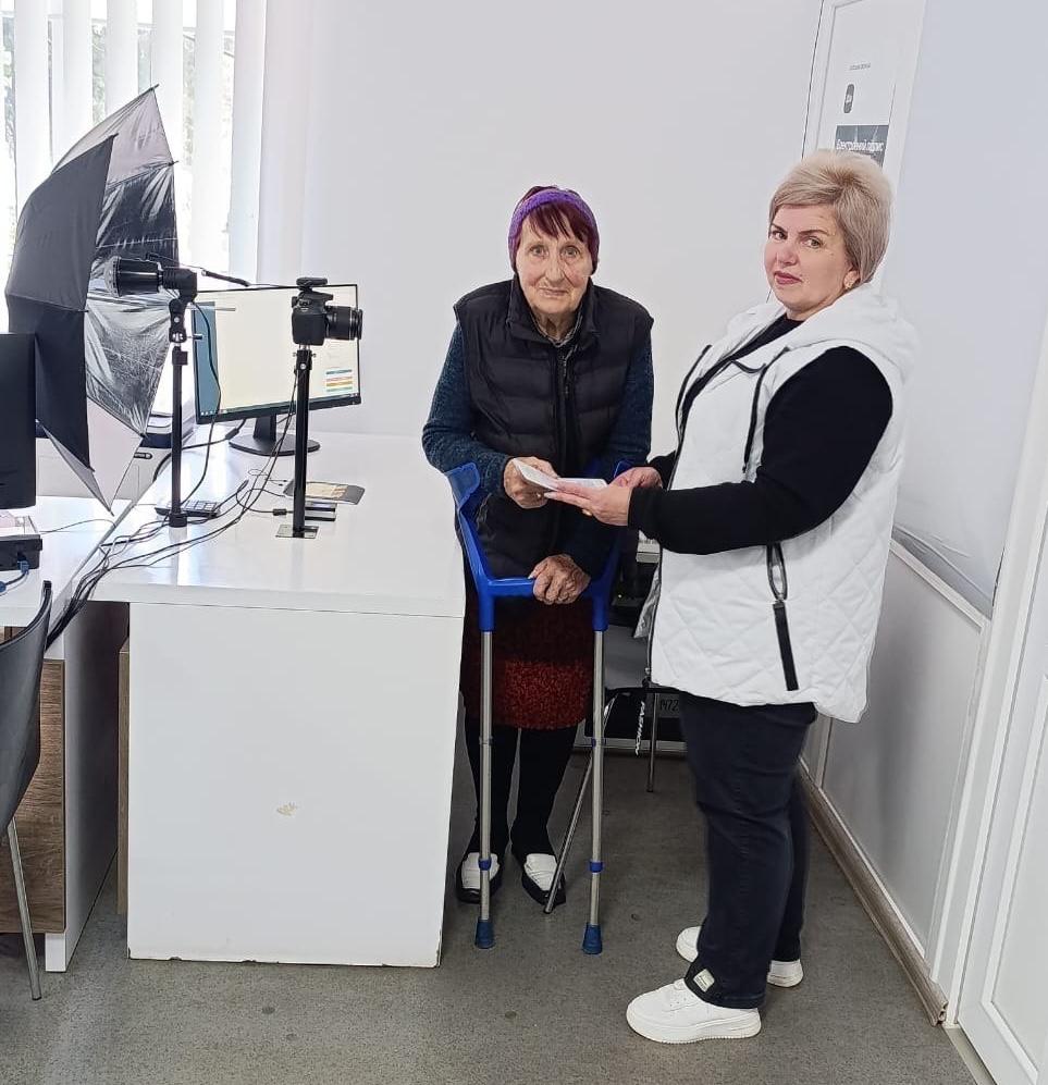 Дві жінки стоять біля білого столу: літня жінка з милицями та інша жінка у білій жилетці, яка передає їй документи, поруч обладнання для фотографування на документи (комп'ютер, камера, парасолька-софтбокс).